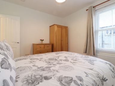 A bedroom with a bed featuring floral bedding a wooden dresser and wardrobe and a window with blinds and curtains at Jacks Cottage in Keswick