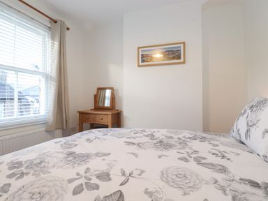 A bedroom with a bed with floral bedding a wooden dressing table with a mirror a window with blinds and curtains and a framed picture on the wall at Jacks Cottage in Keswick