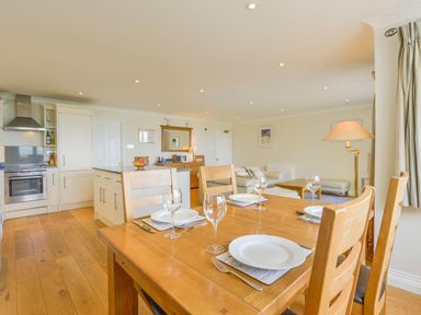 An open kitchen and dining area with a wooden table set for four and a living area with a sofa and floor lamp at 19 Burgh Island Causeway in Bigbury-On-Sea
