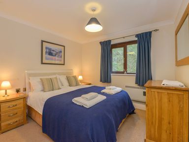 A bedroom with a double bed covered with blue bedding and folded towels on it two wooden bedside tables with lamps a window with blue curtains and a wooden dresser with a mirror at 19 Burgh Island Causeway in Bigbury-On-Sea