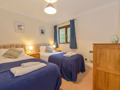 A bedroom with two single beds blue blankets bedside tables lamps a window with blue curtains and wooden dresser at 19 Burgh Island Causeway in Bigbury-On-Sea