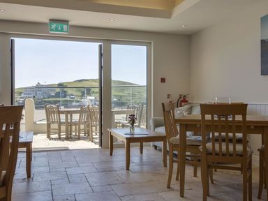 A dining area with wooden tables and chairs next to large glass doors opening to a patio with outdoor seating and a landscape view at 19 Burgh Island Causeway in Bigbury-On-Sea