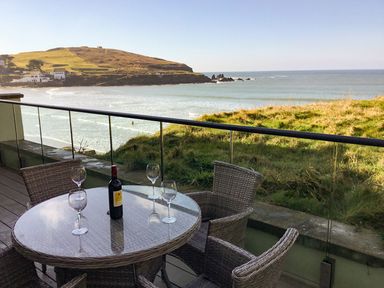A balcony with a round table with a bottle and four wine glasses overlooking the sea and grass at 19 Burgh Island Causeway in Bigbury-On-Sea