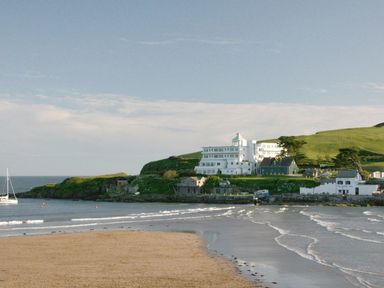 A sandy beach with waves and a white building on a hill near the shoreline at 19 Burgh Island Causeway in Bigbury-On-Sea