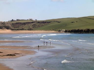 A beach with people carrying surfboards walking into the water and green hills in the background at 19 Burgh Island Causeway in Bigbury-On-Sea