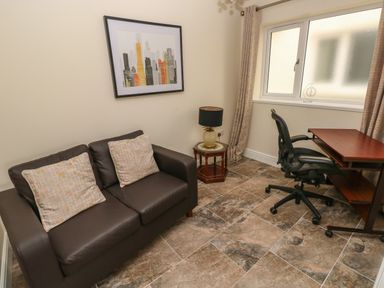 A small room with brown sofa and two cushions a side table with lamp a desk with chair and window at Llygad Y Lleuad Moon in Burry Port