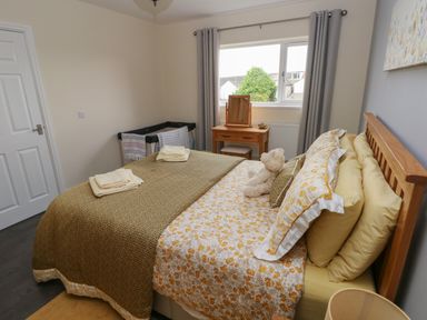 A bedroom with a bed dressed in floral and yellow bedding a teddy bear on the bed a wooden bedside table with a lamp a wooden dressing table and stool in front of a window with grey curtains at Llygad Y Lleuad Moon Burry Port