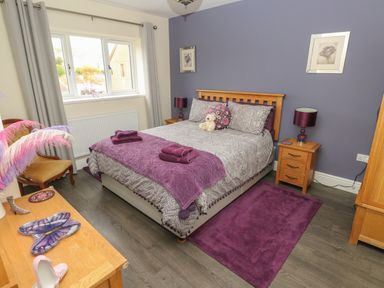 A bedroom with a double bed purple accents wooden furniture and a window with grey curtains at Llygad Y Lleuad ( Moon) in Burry Port