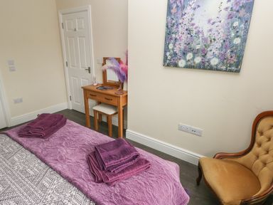 A bedroom with a bed covered with purple towels a wooden dressing table and a chair with a floral painting on the wall at Llygad Y Lleuad Moon in Burry Port