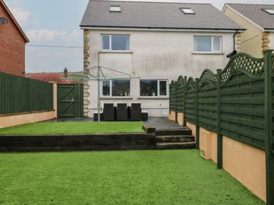 A backyard with green artificial grass, a dark green wooden fence, patio furniture, and a white two-story house at Llygad Y Lleuad Moon in Burry Port