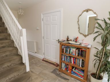 A hallway with carpeted stairs a wooden bookshelf with books a potted plant and a wall mirror at Llygad Yr Haul Sun in Burry Port