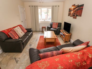 A living room with black leather sofas red cushions a wooden coffee table and a TV on a wooden stand at Llygad Yr Haul Sun in Burry Port