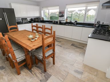 A kitchen with a wooden dining table and chairs white cabinets black countertops and windows showing a green fence at Llygad Yr Haul Sun in Burry Port