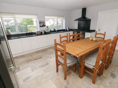 A kitchen with wooden dining table and chairs near windows with white cabinets and black countertops at Llygad Yr Haul Sun in Burry Port