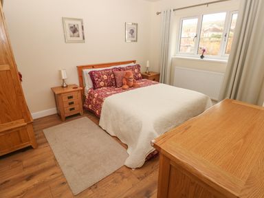 A bedroom with a double bed with floral bedding and a teddy bear bedside tables with lamps wooden furniture and a window with curtains at Llygad Yr Haul Sun in Burry Port