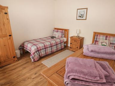 A bedroom with two single beds with plaid bedding a wooden wardrobe a bedside table with a lamp and folded towels on a wooden dresser at Llygad Yr Haul (Sun) in Burry Port