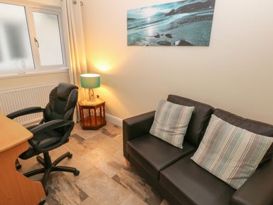 A room with a black leather sofa with two striped pillows a wooden desk with a black office chair a wooden side table with a lamp and a window with closed curtains at Llygad Yr Haul Sun in Burry Port