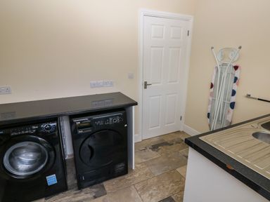 A utility room with washing machine and dryer under a countertop an ironing board by the door and a sink in the corner at Llygad Yr Haul Sun in Burry Port