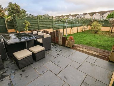 A backyard patio with outdoor dining furniture next to a grass lawn and green wooden fence at Llygad Yr Haul Sun in Burry Port