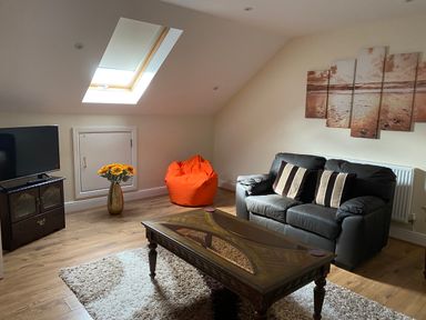 A living room with a black leather sofa, wooden coffee table, orange bean bag, vase with sunflowers, and TV at Llygad Yr Haul Sun in Burry Port