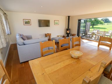 A living room with wooden dining table and chairs a gray sofa a wall-mounted TV a stove and an open door leading to an outdoor seating area at Little Owl Barn in Upton Snodsbury