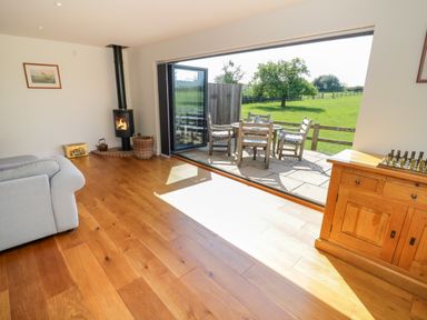A living room with wooden floor a wood stove a gray sofa and a wooden cabinet opening onto a patio with outdoor furniture and green fields at Little Owl Barn in Upton Snodsbury