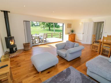 A living room with a stove, wooden floor, gray armchair, ottoman, and large open window to a patio at Little Owl Barn in Upton Snodsbury