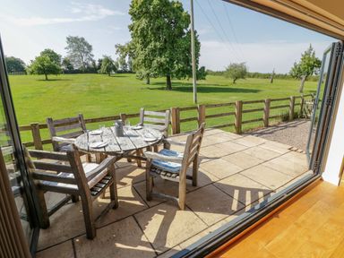 An outdoor patio with a round wooden table and chairs set with plates and glasses overlooking a green field at Little Owl Barn in Upton Snodsbury