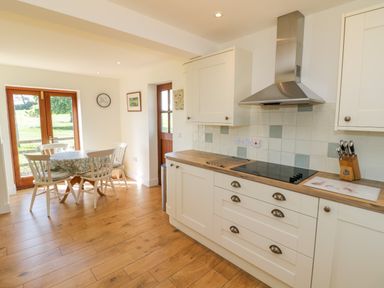 A kitchen area with white cabinets a stove and a dining table with four chairs near wooden doors at Little Owl Barn in Upton Snodsbury