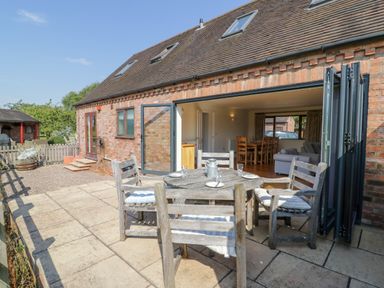 A patio with a wooden table and chairs outside a brick house with open folding doors showing a dining and living area at Little Owl Barn in Upton Snodsbury