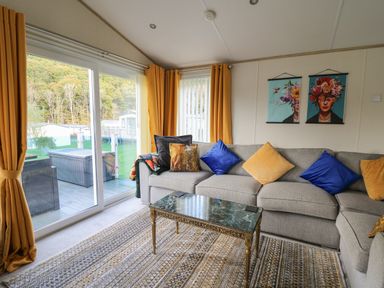 A living room with a gray sectional sofa with blue and yellow pillows a marble top coffee table orange curtains and sliding glass doors at 95 Hafan Taffy in Gilfachrheda near Llanarth