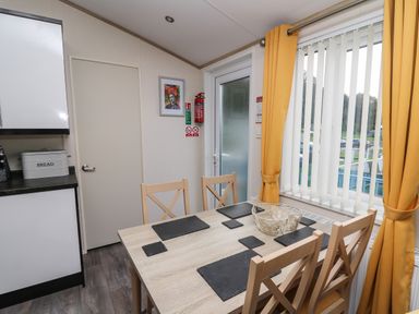 A dining area with a wooden table and four chairs near a window with yellow curtains at 95 Hafan Taffy in Gilfachrheda near Llanarth
