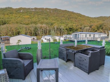 A patio with black wicker chairs and table overlooking a lawn with mobile homes and a forested hill at 95 Hafan Taffy in Gilfachrheda near Llanarth