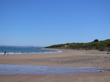 A sandy beach with a few people walking near the water and green hills in the background at 95 Hafan Taffy in Gilfachrheda near Llanarth