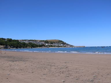A beach with sand and scattered rocks by the sea with boats and houses on a hill in the background at 95 Hafan Taffy in Gilfachrheda near Llanarth