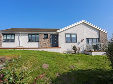 A single story house with white walls stone accents and a grass lawn at Corrie Knowe in Trearddur Bay