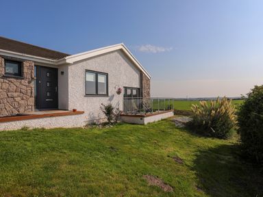 A house exterior with stone and white walls a black door and a glass balcony overlooking a grassy field at Corrie Knowe in Trearddur Bay