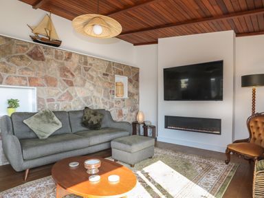 A living room with a gray sofa and ottoman a wooden coffee table a stone wall and a mounted TV with a fireplace below at Corrie Knowe in Trearddur Bay