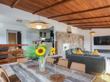 A dining table with sunflowers in a vase and chairs next to a living area with sofas and a wall-mounted tv at Corrie Knowe in Trearddur Bay