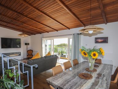 A living room with a grey sofa yellow pillows a wall-mounted tv a fireplace and a dining table with sunflowers at Corrie Knowe in Trearddur Bay