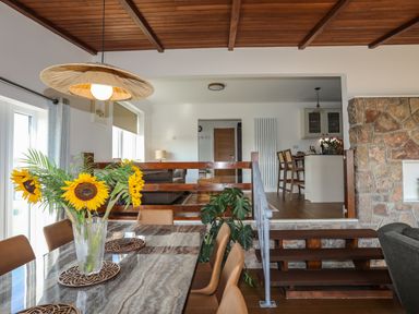 A dining area with a marble table and sunflowers in a vase overlooking a living room and kitchen separated by wooden railing and steps at Corrie Knowe in Trearddur Bay