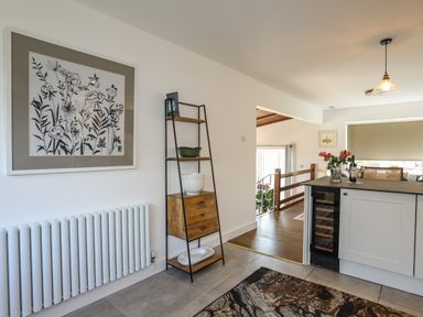 A kitchen area with a shelf unit holding bowls and plates next to a radiator and kitchen counter with flowers in a vase at Corrie Knowe in Trearddur Bay