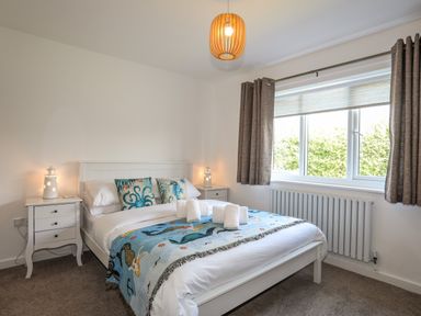 A bedroom with a bed with ocean-themed pillows and blanket bedside tables with lamps and a window with curtains at Corrie Knowe in Trearddur Bay