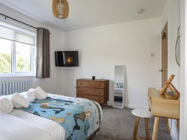 A bedroom with a bed covered by a mermaid blanket three rolled towels on the bed a wooden chest of drawers a wall-mounted television a full-length mirror and a wooden desk with a stool at Corrie Knowe in Trearddur Bay