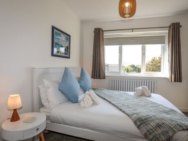 A bedroom with a bed two blue pillows bedside table lamp and window with curtains at Corrie Knowe in Trearddur Bay