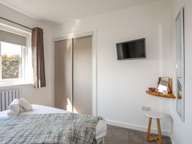 A bedroom with a bed towel by the window curtain a wall mounted TV a small wooden shelf with a mirror and a stool at Corrie Knowe in Trearddur Bay