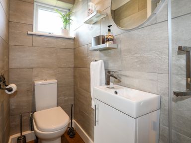A small bathroom with a toilet a sink a round mirror a potted plant on the window ledge and toiletries on wall shelves at Corrie Knowe in Trearddur Bay