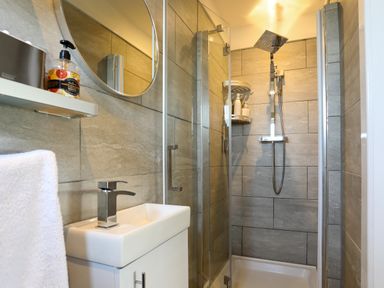 A bathroom with a rectangular sink and faucet a round mirror and a glass shower enclosure with a showerhead and shelves holding toiletries at Corrie Knowe in Trearddur Bay