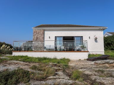 A single-story house with a stone chimney and glass balcony overlooking rocky ground at Corrie Knowe in Trearddur Bay