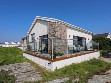 A house with a stone and white exterior and a glass balcony with chairs on a grassy and rocky landscape at Corrie Knowe in Trearddur Bay